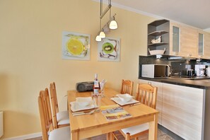 A dining area with a wooden table, chairs, and a radio. There are two paintings on the wall, a kitchen with a microwave and cabinets, and a shelf with decorative items.