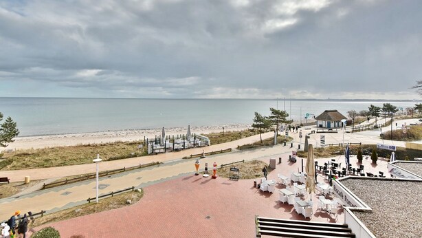 A beachside promenade with outdoor seating, a thatched-roof structure, and a view of the sea.
