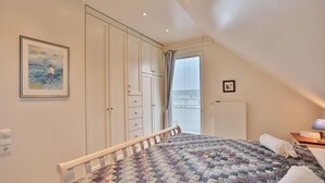 Een slaapkamer met een bed, een kledingkast, een raam met gordijnen, een radiator en een ingelijste foto aan de muur.