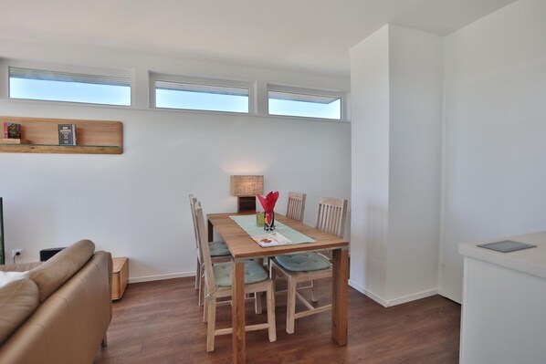 Un comedor con una mesa de madera, cuatro sillas y una lámpara de mesa pequeña. Hay dos ventanas sobre la mesa y una estantería con libros a la izquierda.