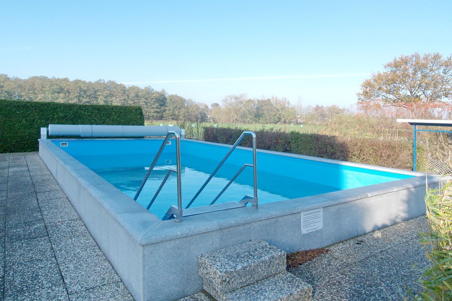A rectangular swimming pool with metal ladders, surrounded by stone tiles and a hedge.