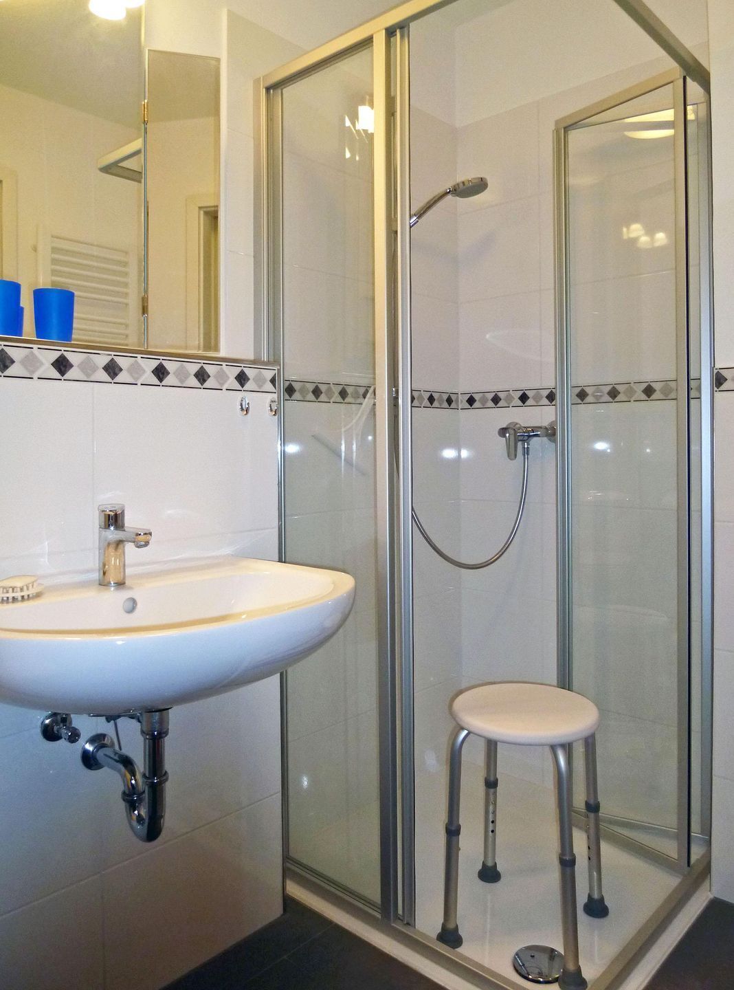 Une salle de bain avec une douche à parois de verre, un lavabo avec un miroir et un petit tabouret.