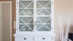 A white cabinet with glass doors, containing bowls, glasses, and a heart-shaped decoration. - Strandglück Apartment 8A StrandGutFinder.de - Strandglück Haus A (Scharbeutz)