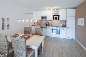 Ett modernt kök med vita skåp, en mikrovågsugn, en ugn och en matplats med flätade stolar och ett vitt bord.