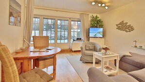 A cozy living room with a wooden dining table, a comfortable sofa, a television, and a large window with curtains.