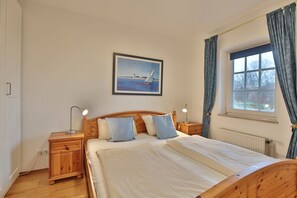 A bedroom with a wooden bed, two bedside tables with lamps, a window with curtains, and a painting on the wall. - Seepark Appartement 13 - Seepark (Scharbeutz)