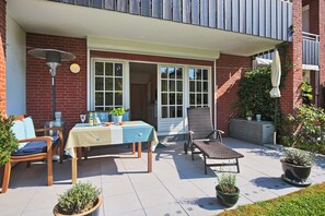 Eine Terrasse im Freien mit Tisch und Stühlen, einem Sessel, Topfpflanzen und einem Gebäude mit Backsteinfassade und Balkon.