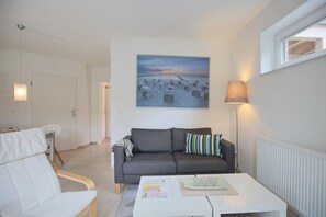 A modern living room with a grey sofa, a white armchair, a coffee table, and a painting of a sunset over a beach. - Haus Küstenkinder Marie Apartment 1 - Haus Küstenkinder (Scharbeutz)