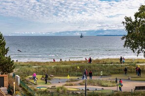 A coastal area with a golf course, a body of water, and a sailboat in the distance. - Seetempel Apartment 42 - Seetempel (Scharbeutz)