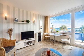 Een moderne woonkamer met een flatscreen televisie, een eethoek met een tafel en stoelen, een groot raam met uitzicht op zee en een balkon met een hekwerk.