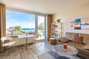 Een moderne woonkamer met uitzicht op zee, een eethoek met tafel en stoelen, een boekenkast en een bank.