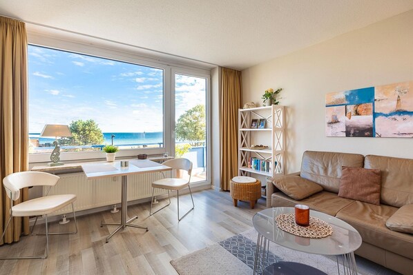 A modern living room with a view of the sea, a dining area with a table and chairs, a bookshelf, and a sofa.