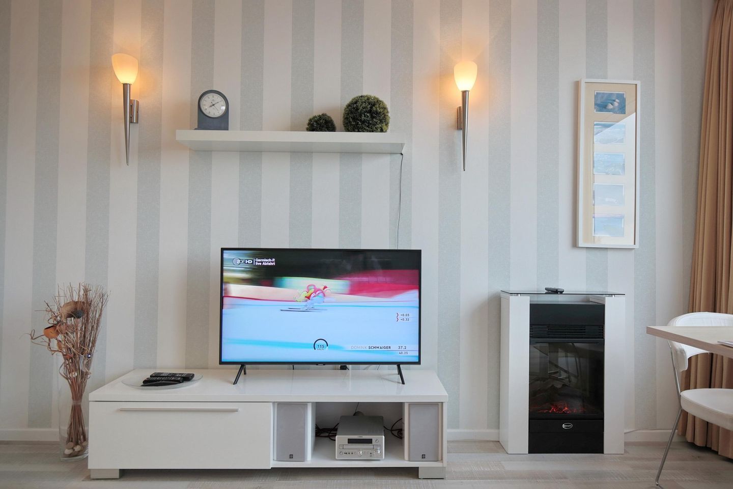 A modern living room with a flat-screen TV on a white entertainment unit, a fireplace, and a striped wall.