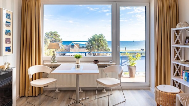 Een kamer met een groot raam dat uitkijkt op een strand, een tafel met stoelen, een lamp en een boekenkast.