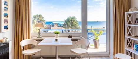 A room with a large window overlooking a beach, a table with chairs, a lamp, and a bookshelf.
