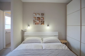 A bedroom with a bed, two bedside tables, a wall-mounted shelf with decorative items, and a window.