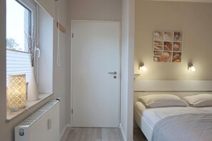 A bedroom with a bed, a window with a view, a door, and a wall with a light fixture and a framed picture. - Seetempel Apartment 39 - Seetempel (Scharbeutz)