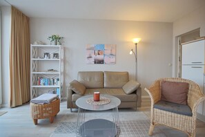 Un salon comprenant un canapé, une table basse, un fauteuil en osier, une bibliothèque et un tableau accroché au mur.