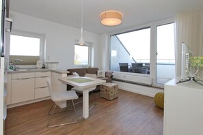 A modern, well-lit room with a kitchen, dining area, and large windows offering a view. - Seetempel 45 Apartment - Seetempel (Scharbeutz)