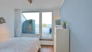 Een slaapkamer met een bed, een televisie op een kast, een raam met uitzicht op zee en een plafondlamp.