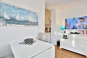 Un salon moderne avec un canapé blanc, une table à manger agrémentée d’un panier, un téléviseur fixé au mur et un tableau représentant des voiliers.