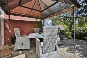 Un patio couvert aménagé avec des meubles en osier et une table, agrémenté d’un mur en briques et d’une végétation luxuriante en arrière-plan.