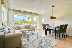 A modern living room with a sofa, coffee table, dining table, and chairs. There is a large window providing a view of the outside. - Feriendomizil Neue Bergstrasse 11 Appartement 4 - Feriendomizil Neue Bergstrasse (Scharbeutz)