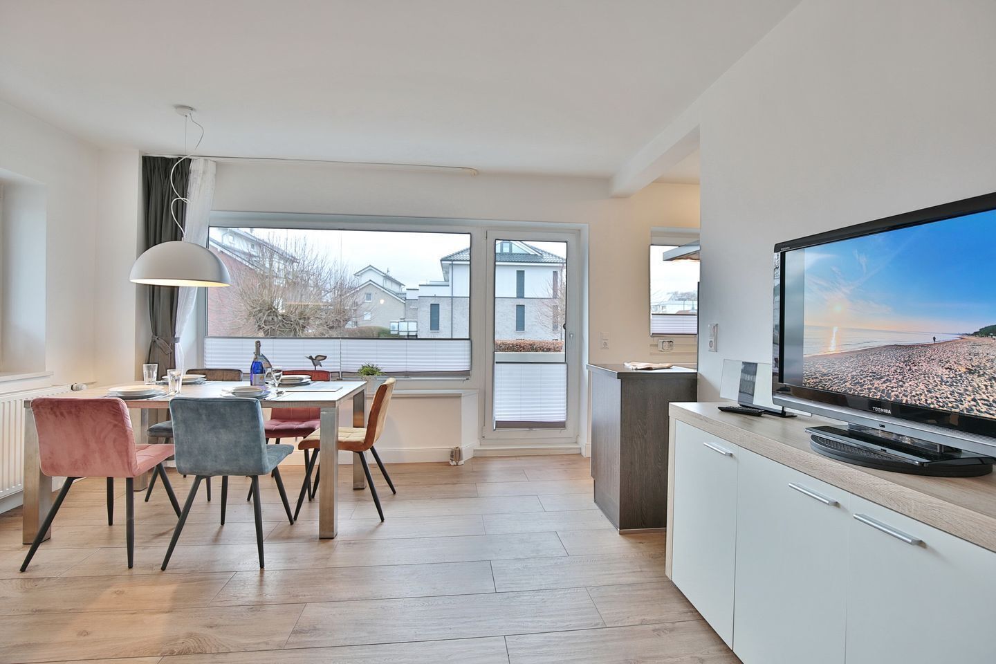 A modern kitchen with a dining area, large windows, and a flat-screen TV.