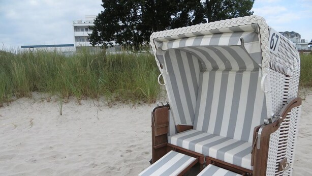 A beach chair with a striped cushion and a number 67 sign, set on sandy ground with grass and a building in the background.