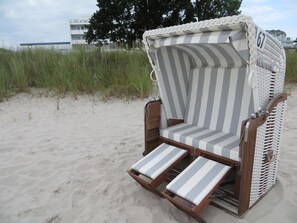 A beach chair with a striped cushion and a number 67 sign, set on sandy ground with grass and a building in the background. - Vacation Rental Neue Bergstrasse 11, Apartment 8 - Vacation Rental Neue Bergstrasse (Scharbeutz)