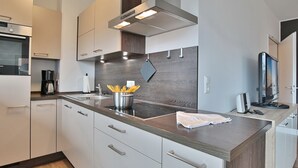 A modern kitchen with white cabinets, a stainless steel range hood, a built-in oven, a coffee maker, and a TV on a counter.
