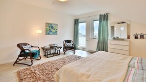 Un dormitorio con cama, dos sillas, una mesa, una lámpara de pie, un cuadro, un aparador y una ventana con cortinas.