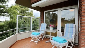 Un balcón con dos sillones blancos acolchados, una mesita con un florero y una puerta de cristal que da a vistas de árboles y un cielo nublado.