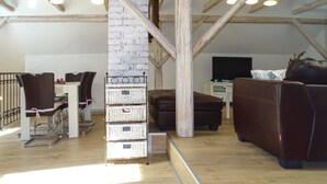 Een loft-achtige woonruimte met zichtbare houten balken, een witte bakstenen accentmuur, een bank, een eettafel met stoelen en een televisie.