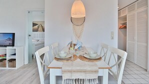 A dining area with a white table set for four, a hanging light fixture, and a television in the background.
