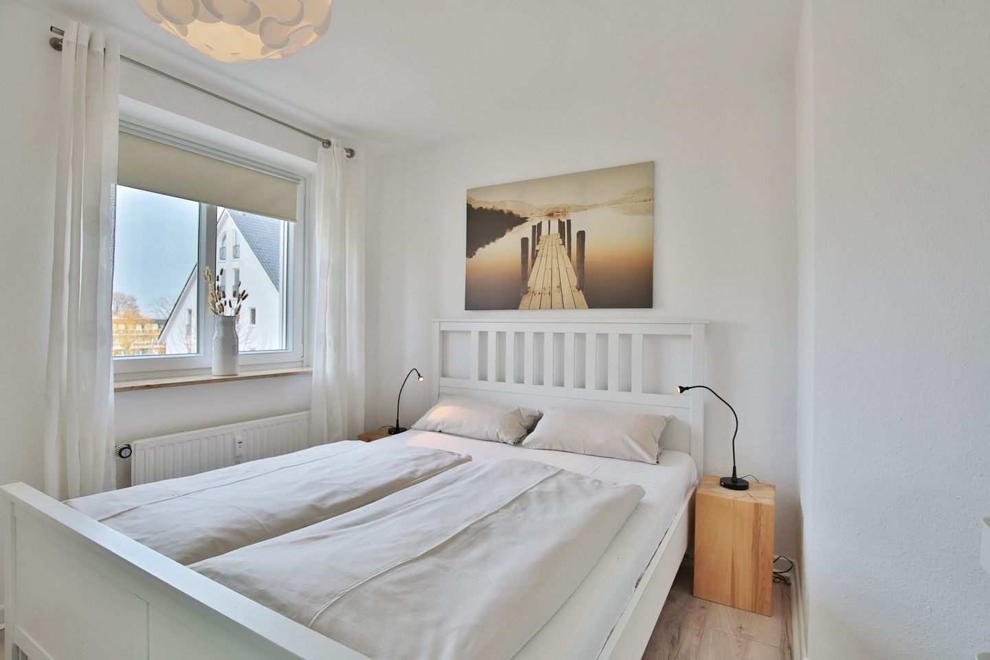 Um quarto com cama, duas mesas de cabeceira, uma janela com cortinas, um radiador e um quadro na parede.