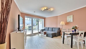 A modern living room with a grey sectional sofa, a dining table with white chairs, and a kitchen area with a white cabinet and a stove. - Beach villa apartment 6 - Beach villa (Scharbeutz)