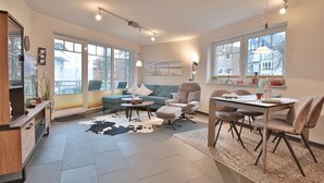 A modern living room with a sofa, armchairs, a dining table, and a large window. - Beach villa apartment 14 - Beach villa (Scharbeutz)