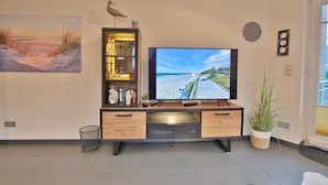 A modern living room with a large flat-screen TV, a bookshelf with decorative items, and a painting of a beach scene. - Beach villa apartment 14 - Beach villa (Scharbeutz)