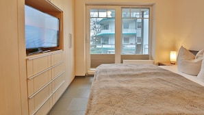 A hotel room with a bed, a TV on a wooden cabinet, a balcony with a view of buildings, and a nightstand with a lamp. - Beach villa apartment 14 - Beach villa (Scharbeutz)