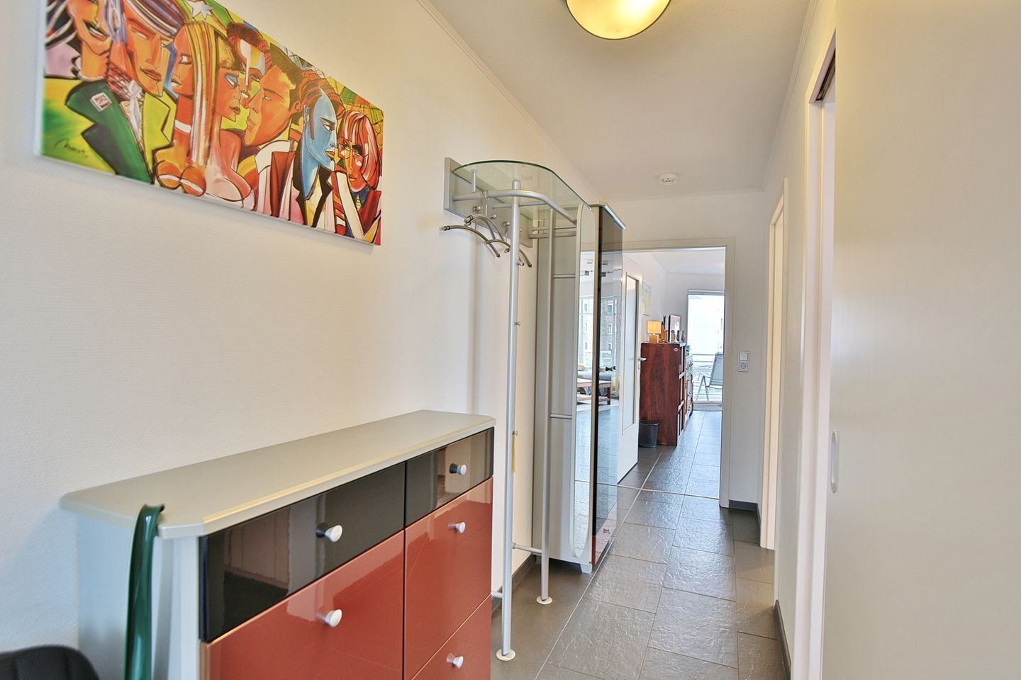 Ein Flur mit einer Glastür zu einem Raum, ein farbiges abstraktes Gemälde an der Wand und ein modernes Schrank mit Schubladen.