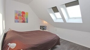 A bedroom with a sloped ceiling, a bed with a brown quilt, a desk with a computer, and a colorful abstract painting.