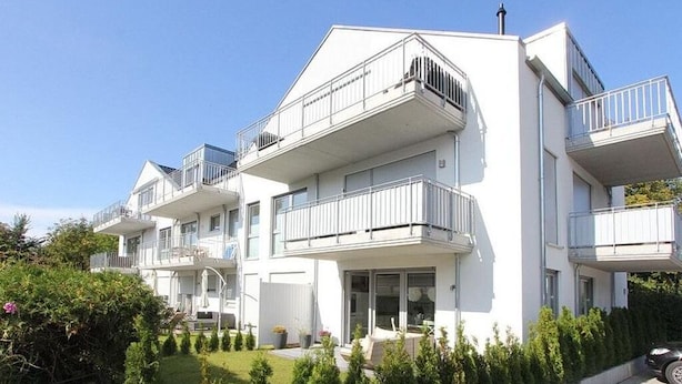 A modern multi-story residential building with balconies, a well-maintained garden, and a paved driveway.