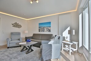 Una sala moderna con un sofá gris, dos sillones, una mesita de centro y una mesita auxiliar con una lámpara. Hay un cuadro de una escena de playa en la pared y un adorno decorativo sobre el sofá.