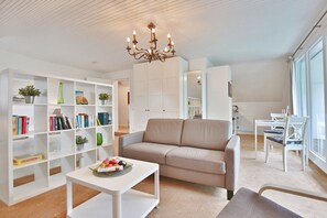 Una sala moderna con un sofá beige, estanterías blancas con libros y plantas, una mesa de centro blanca y una araña.
