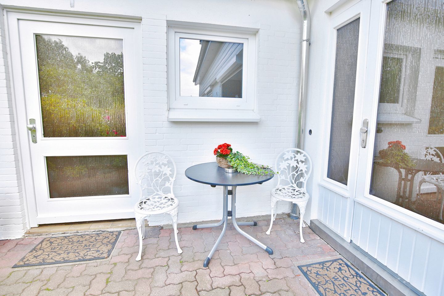 Un mur extérieur blanc avec une fenêtre et une porte, une petite table ronde avec des chaises et un paillasson.