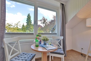 A room with a round table, two white chairs, a window with a view of trees, and a radiator.