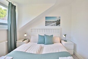 A bedroom with a white headboard, two bedside tables with lamps, a bed with white and teal bedding, and a picture of a beach scene on the wall. - Apartment house Linda-Lu Apartment 9 - Apartment house Linda-Lu (Scharbeutz)