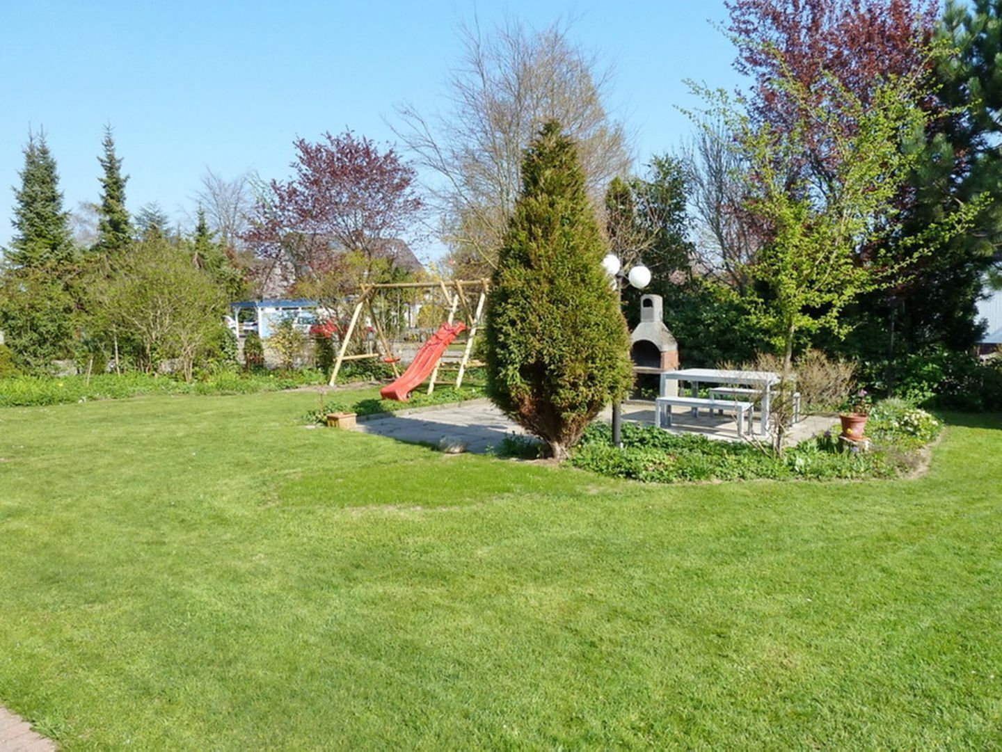 A well-maintained garden with a wooden swing set, a small brick structure, and a variety of trees and shrubs.