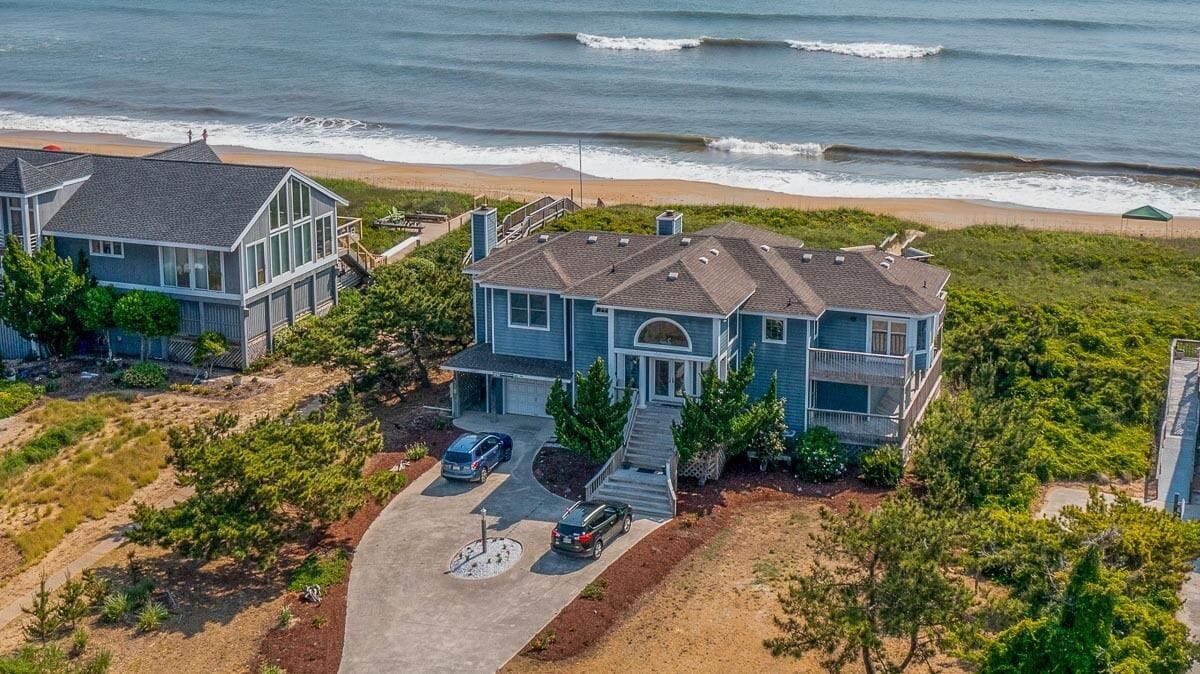 A coastal house with a large driveway, two cars parked, and a view of the ocean.